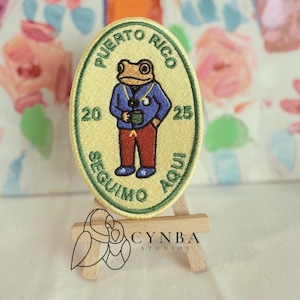 May include: Embroidered patch with a frog wearing a blue jacket, red pants, and holding a mug. The patch reads "Puerto Rico" and "Seguimo Aqui" in green lettering, with the numbers "20" and "25". The patch is oval-shaped with a yellow background.