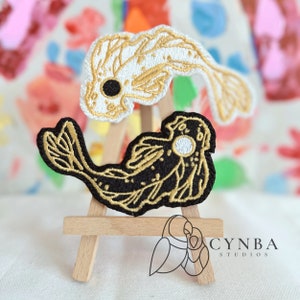 May include: Two embroidered iron-on patches of koi fish, one black with gold accents and the other white with gold accents. The patches are displayed on a small wooden easel.