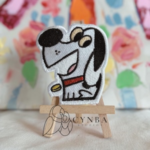 May include: Embroidered patch featuring a cartoon dog with a red collar and a gold coin. The dog is white with black accents and a smiling expression. The patch is displayed on a small wooden easel.