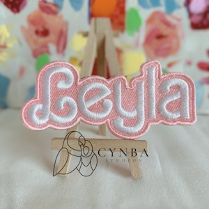 May include: Pink and white embroidered patch with the name "Beyla" in a cursive font. The patch is displayed on a small wooden easel.