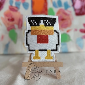 May include: Embroidered patch featuring a pixelated chicken wearing black sunglasses. The chicken has a white body, yellow feet, an orange and yellow beak, and a red chest. The patch is displayed on a small wooden easel. The text "CYNBA STUDIOS" is visible.
