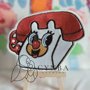 May include: White embroidered patch of a cartoon phone with a red receiver. The phone has a smiling face with blue eyes and a red nose. The patch is on a small wooden easel with the text "CYNBA STUDIOS" below.
