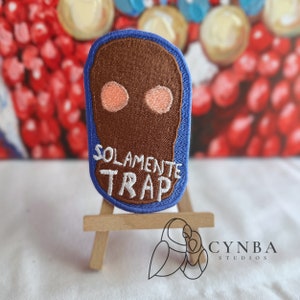May include: A blue and brown embroidered patch with the text "Solamente Trap" in white. The patch is on a small wooden easel.