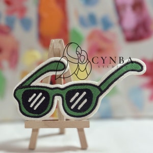 May include: Green embroidered sunglasses patch with white stitching. The patch is displayed on a small wooden easel.
