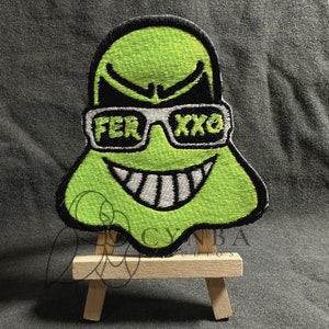 May include: Green ghost-shaped embroidered patch with a black outline, wearing sunglasses that say "FER XXO" and a white toothy grin. The patch is displayed on a small wooden easel.
