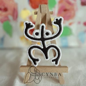 May include: An embroidered patch featuring a black Taino Coqui symbol with a thick white border. The patch is displayed on a small wooden easel that has the text CYNBA STUDIOS and a line art logo of two birds printed on the bottom crossbar.