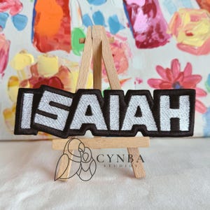 May include: Embroidered patch with the name "ISAIAH" in white block letters on a black background. The patch is displayed on a small wooden easel in front of a colorful abstract painting. The brand logo "CYNBA STUDIOS" is at the bottom.