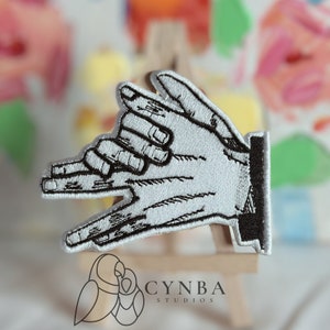 May include: A white embroidered patch depicting two hands forming a shadow puppet of a dog. The patch is on a wooden stand with the text "CYNBA STUDIOS" below.