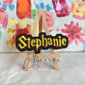 May include: A black embroidered patch with the name "Stephanie" in yellow, displayed on a small wooden easel. The patch has a decorative border and is set against a colorful abstract background. The patch is a personalized item.