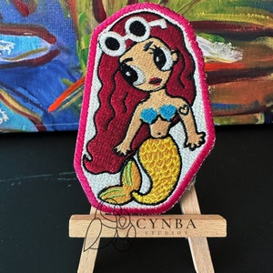 May include: Embroidered patch of a cartoon mermaid with red hair, wearing round sunglasses and a blue bikini top. The mermaid has a yellow tail with green accents. The patch is surrounded by a pink border.