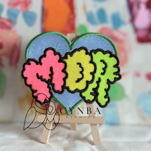 May include: A heart-shaped patch with the word "Mop" in colorful, puffy letters. The letters are pink, green, and yellow, outlined in black. The patch is on a blue background and is mounted on a small wooden easel.