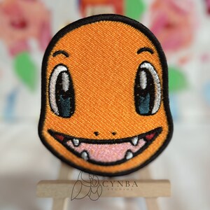 May include: Orange and black embroidered patch of a cartoon character with large eyes and a wide smile. The character has a small nose and a pointed chin.