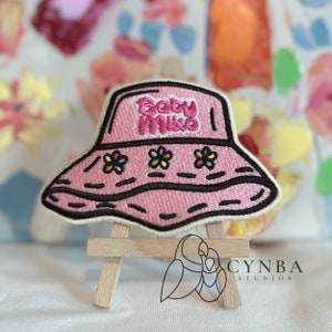 May include: Pink embroidered patch of a bucket hat with the text "Baby Mike" on the crown. The hat has a black outline and three embroidered flowers on the brim.