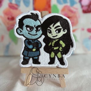 May include: Embroidered patch featuring two cartoon characters. One character is blue with black hair and a stern expression, wearing a blue outfit. The other character is green with black hair and a confident expression, wearing a green and black outfit.