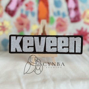 May include: A black and white embroidered patch with the word "keveen" in white lettering.