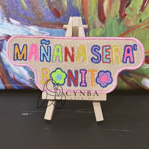 May include: A pink and beige embroidered patch with the text "Mañana Sera Bonito" in colorful letters. The patch is displayed on a small wooden easel.