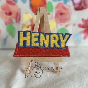 May include: A rectangular patch with the name "HENRY" embroidered in yellow letters with a blue outline, set against a red background. The patch is displayed on a miniature wooden easel, with the "CYNBA STUDIOS" logo visible.