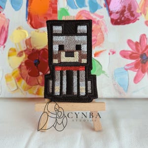 May include: Embroidered patch featuring a pixelated dog design with a black outline, gray and tan body, and a red collar. The patch is displayed on a small wooden easel. The text "CYNBA STUDIOS" is visible.