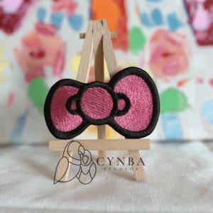 May include: A pink embroidered bow patch with a black outline, displayed on a small wooden easel. The bow has a textured appearance. The background is a soft, floral pattern. The patch is a decorative item.