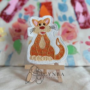 May include: Embroidered patch featuring a cartoon cat in shades of orange, tan, and white. The cat is sitting with its tail curled. The patch has a white border and is displayed on a small wooden easel. The patch has the text "CYNBA STUDIOS".
