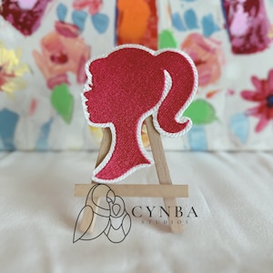 May include: A red embroidered patch in the shape of a woman's profile with a ponytail, outlined in white. The patch is displayed on a small wooden easel. The logo "CYNBA STUDIOS" is visible in the lower right corner.