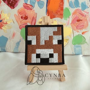 May include: A square embroidered patch featuring a pixelated cow face design in shades of brown, white, and black. The patch is displayed on a small wooden easel. The embroidery has a distinct grid-like pattern with sharp, blocky edges.