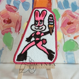 May include: Pink bunny rabbit wearing a black and white swimsuit holding a multi-colored ice cream cone. The bunny is embroidered on a white patch with a pink border.