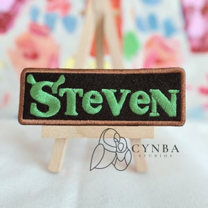May include: Embroidered patch with the name "Steven" in green letters on a black background. The patch has a brown border and is displayed on a small wooden easel. The logo "CYNBA STUDIOS" is visible below the patch.
