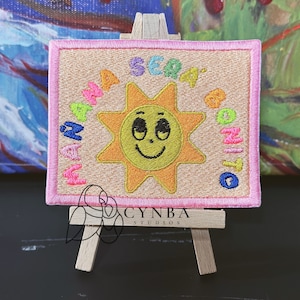 May include: A pink and beige embroidered patch with a smiling sun and the text "WAP SE4 DA ON1T0" around it.