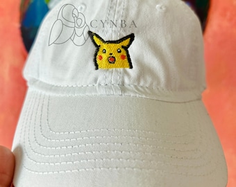 May include: White baseball cap with an embroidered yellow Pikachu meme image. The text "CYNBA STUDIOS" is embroidered on the front of the cap.