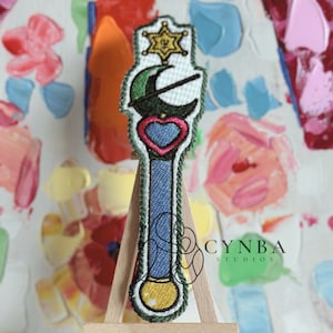 May include: A green, blue, and gold embroidered patch of a magical wand with a star, crescent moon, and heart. The wand is on a small wooden easel.