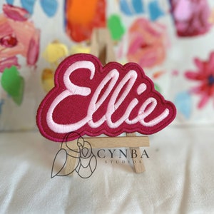May include: Pink and red embroidered patch with the name "Ellie" in a cursive font. The patch is displayed on a small wooden easel.