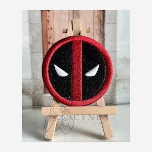 May include: Red and black embroidered patch with a white outline of Deadpool's mask. The patch is displayed on a small wooden easel.