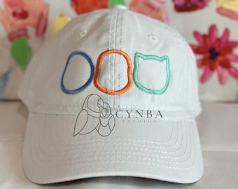 May include: White baseball cap with a blue, orange, and green embroidered design of three circles and a cat head. The text "CYNBA STUDIOS" is embroidered on the front of the cap.