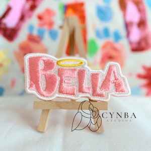 May include: A pink embroidered patch with the name "BELLA" in a white outline. The letters have a halo above them. The patch is displayed on a small wooden easel.
