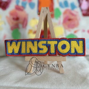 May include: Embroidered patch with the name "WINSTON" in yellow, outlined in blue, and set against a red background. The patch is displayed on a small wooden easel, with the "CYNBA STUDIOS" logo visible below.