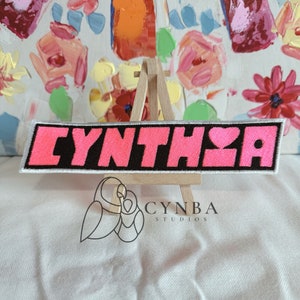 May include: A pink and black embroidered patch with the name "CYNTHIA" and a heart in the middle. The patch is on a wooden easel.