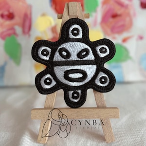 May include: A black and white embroidered patch featuring a stylized sun-like symbol with a central circular face and six radiating petal-like extensions. The patch is displayed on a small wooden easel with the text CYNBA STUDIOS printed below.