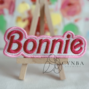 May include: A pink and white embroidered patch with the name "Bonnie" in a bold, cursive font. The patch is displayed on a small wooden easel.