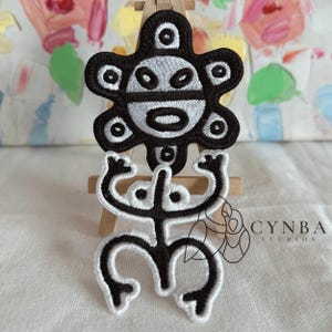 May include: Two embroidered patches with black and white designs. The top patch is a stylized sun shape with a face. The bottom patch depicts a figure with arms raised. The patches are displayed on a small wooden easel.