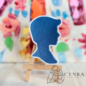 May include: A blue fabric silhouette of a person's head, displayed on a small wooden easel. The silhouette is a simple outline of a person's head and shoulders, with no facial features.