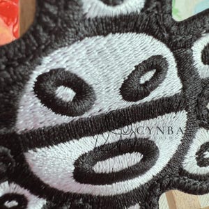 May include: Close-up of a black and white embroidered patch. The design features a repeating pattern of oval shapes with a central eye-like detail. The patch is intricately stitched with a textured appearance.