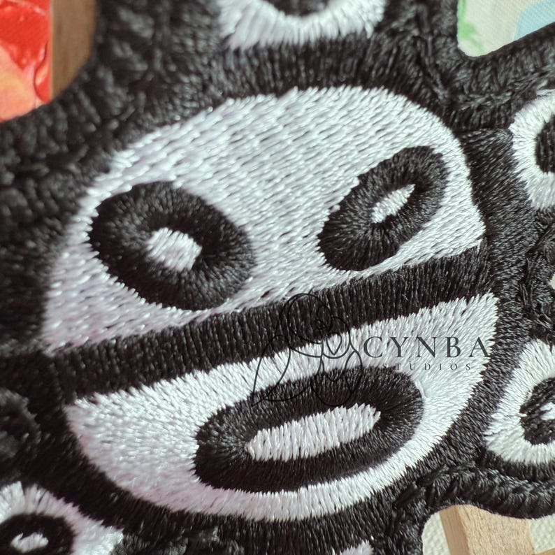 May include: Close-up of a black and white embroidered patch. The design features a repeating pattern of oval shapes with a central eye-like detail. The patch is intricately stitched with a textured appearance.