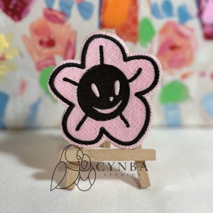 May include: Pink embroidered flower patch with a black smiling face in the center. The patch is displayed on a small wooden easel.