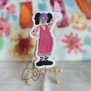 May include: A colorful embroidered patch featuring a smiling clown in a pink jumpsuit with a purple hat and black shoes. The patch is on a small wooden easel with the text "CYNBA STUDIOS".