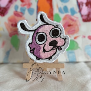 May include: Embroidered patch featuring Courage the Cowardly Dog. The patch has a pink body, black eyes and nose, and a white outline. The patch is displayed on a small wooden easel. The Cynba Studios logo is visible.