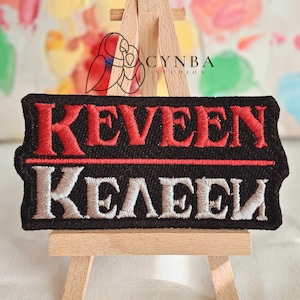May include: A black embroidered patch with the name "KEVEEN" in red thread above "KELEEN" in white thread. The patch is rectangular with rounded corners and is displayed on a small wooden easel.