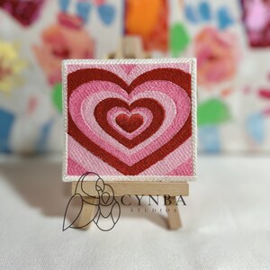 May include: Square embroidered patch featuring a concentric heart design in shades of red and pink. The patch has a white border and is displayed on a miniature wooden easel. The patch is a decorative item.