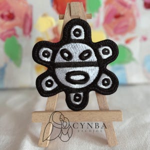 May include: Black and white embroidered patch featuring a stylized sun design. The patch has a black border and a white interior with circular and oval shapes. The patch is displayed on a small wooden easel. The text "CYNBA STUDIOS" is visible.