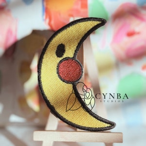 May include: A yellow crescent moon shaped patch with a red circle and a black eye. The patch is embroidered with black thread and has a black outline. The patch is on a wooden easel with the text "CYNBA STUDIOS" in black text.
